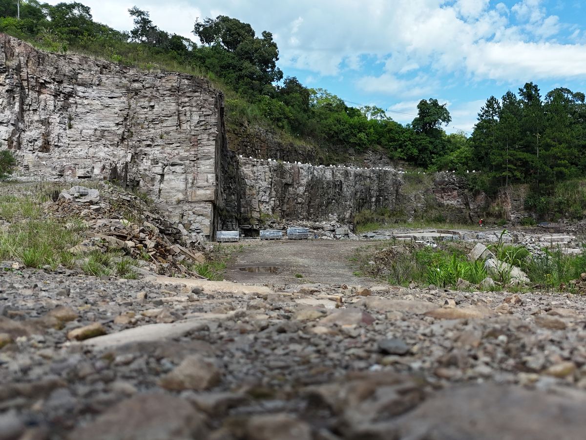 Cava de basalto vista do nível do chão, com parede rochosa estratificada, pallets de pedra talhada e mata nativa ao fundo sob céu azul