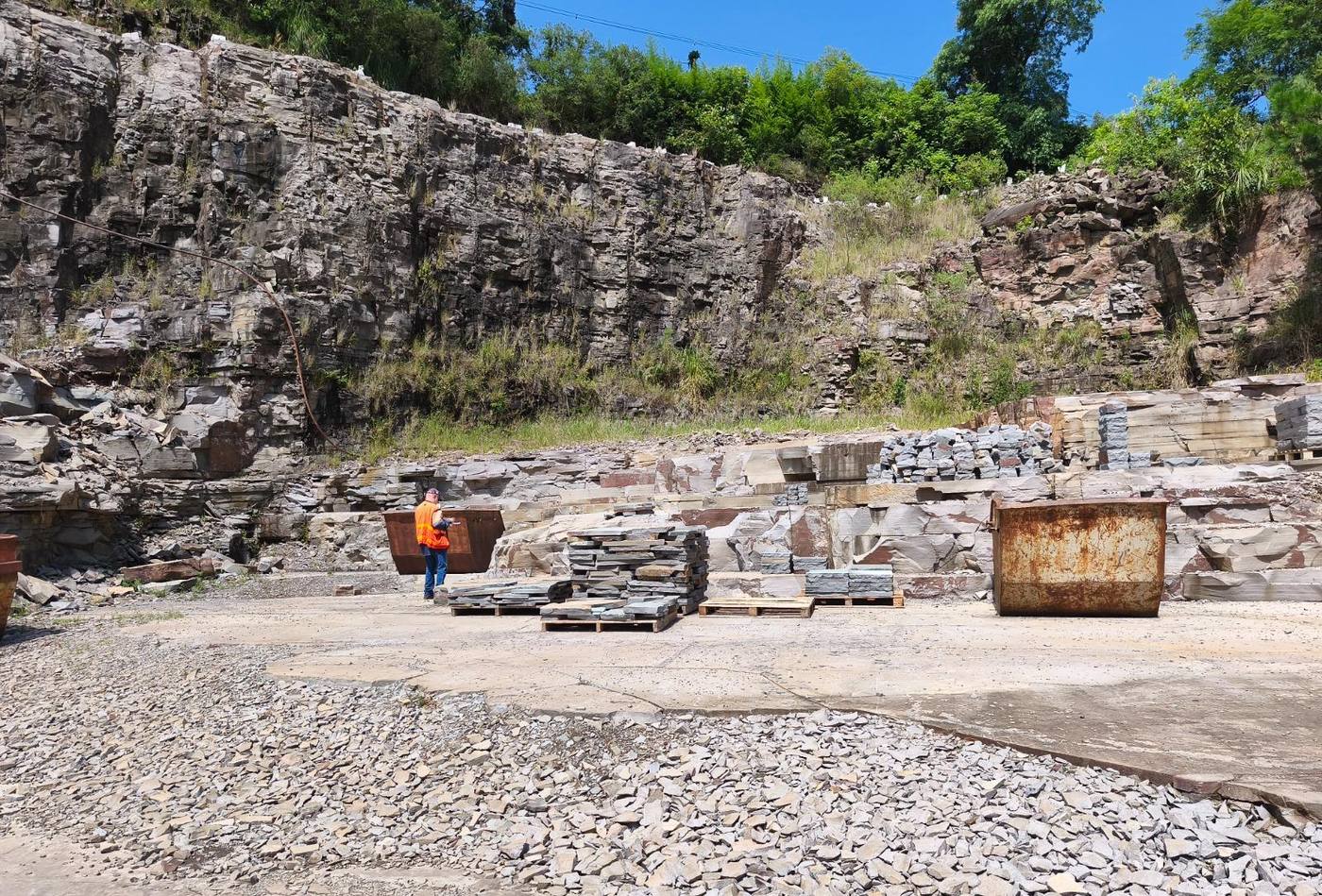 Pátio de pedreira de basalto com pallets de lajes e pedras talhadas, caçambas e um profissional de colete laranja fazendo vistoria diante do paredão