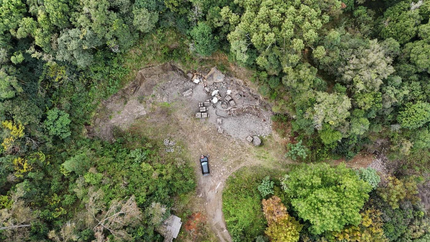 Vista aérea de drone, de cima para baixo, de uma pequena lavra de basalto em clareira cercada por mata nativa densa, com pallets de pedra e um veículo