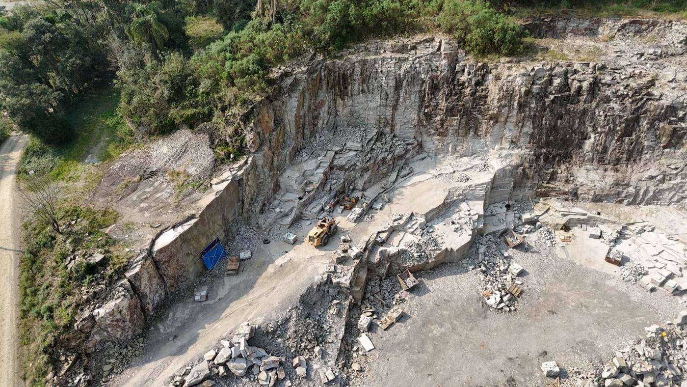 Vista aérea de drone de uma pedreira de basalto em operação, com pá carregadeira, pallets de pedra talhada e paredões rochosos cercados por mata